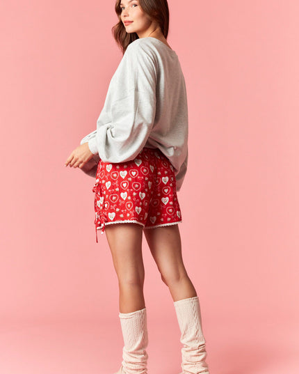 Woman wearing a gray sweatshirt, red shorts with heart patterns, and white socks on a pink background