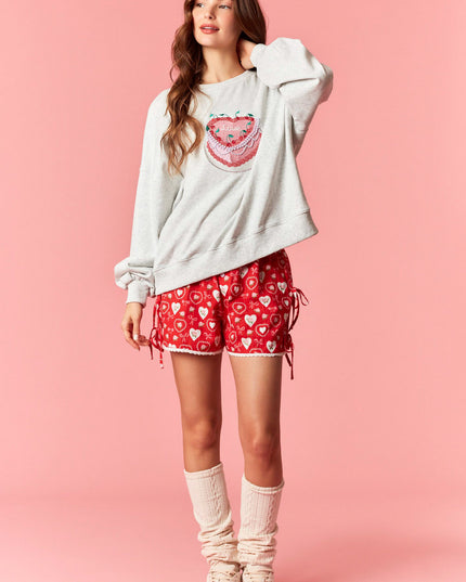 Woman wearing a white sweatshirt with a graphic design and red shorts with heart patterns on a pink background