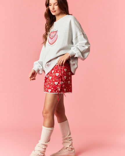 Woman wearing a white sweatshirt with a graphic design and red shorts with heart patterns on a pink background