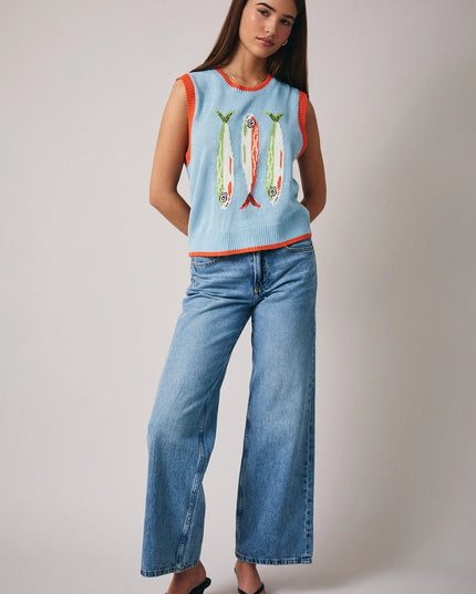 Woman wearing a light blue sleeveless top with fish designs and high-waisted blue jeans on a plain background
