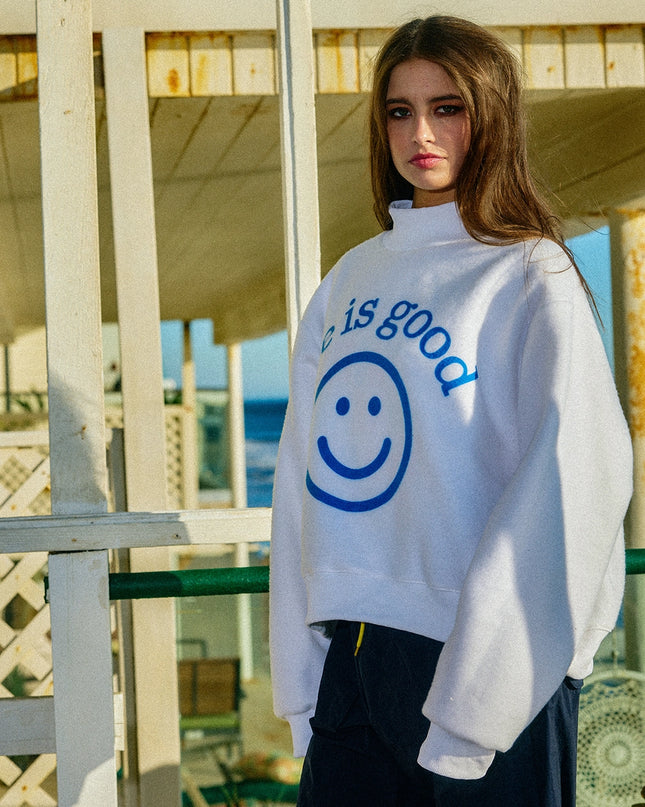 Person wearing a white sweatshirt with a smiley face and 'is good' text, standing outdoors.
