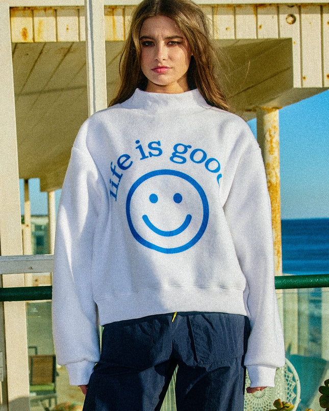 Person wearing a 'life is good' sweatshirt with a smiley face graphic by a beach.
