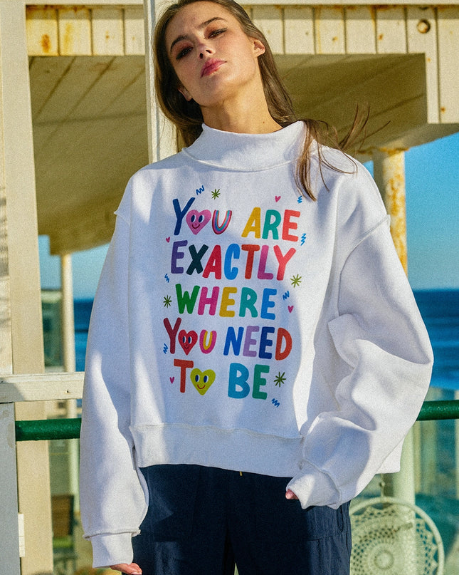Person wearing a white sweatshirt with colorful text by the beach . the sweatshirt says you are exactly where you need to be sweatshirt