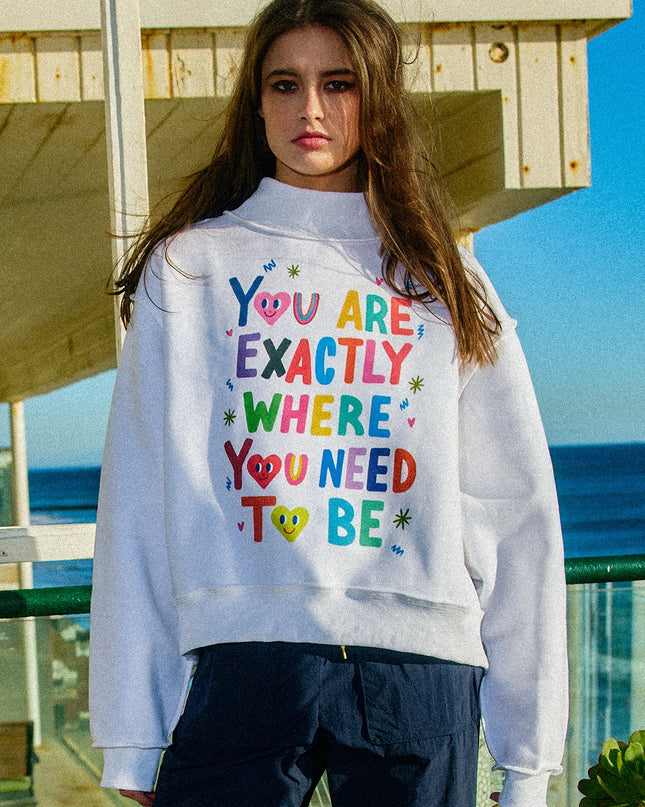 Person wearing a grey sweatshirt with colorful text  that says "you are exactly where you need to be" with doodles and is standing by the ocean.