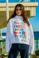 Person wearing a grey sweatshirt with colorful text  that says "you are exactly where you need to be" with doodles and is standing by the ocean.